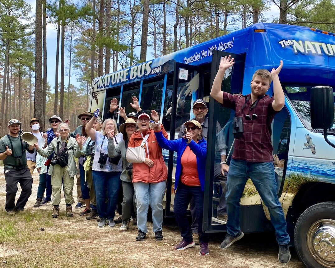 Group of people posing excitedly in front of The Nature Bus with their eco-tour guide in a pine forest.